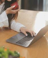 Portrait of pretty freelancer female working typing on laptop computer sitting at table smiling looking to screen in kitchen. Attractive redhead young woman remote working on laptop from home office. photo