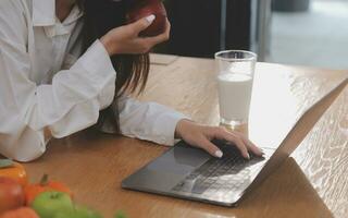 Portrait of pretty freelancer female working typing on laptop computer sitting at table smiling looking to screen in kitchen. Attractive redhead young woman remote working on laptop from home office. photo