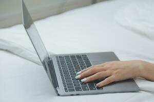 Happy young female in casual clothes smiling and looking at camera while using laptop on bed in bedroom at home photo