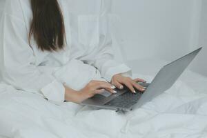 Happy young female in casual clothes smiling and looking at camera while using laptop on bed in bedroom at home photo