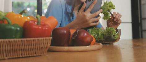 A young woman with a beautiful face in a blue shirt with long hair eating fruit sitting inside the kitchen at home with a laptop and notebook for relaxation, Concept Vacation. photo