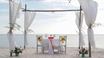 Romantic dinner table set and chair preparation with white theme decoration, white sand, clear cloudy sky, and tranquil, peaceful, beautiful beach view at the seashore. Good place for wedding proposal photo