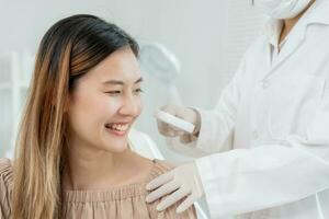 hermosa asiático mujer sonrisa durante recibir piel cuidado. especialista de piel utilizar aumentador vaso Mira que produce picor piel desde piel alérgico, esteroide alergia, sensible piel, químico alergia, erupción, dermatitis. foto