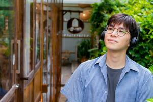 retrato de asiático joven adulto en azul camisa, vistiendo los anteojos, participación auriculares y en pie en frente de el puerta de café comercio, concepto para estilo de vida de adolescentes alrededor el mundo. foto