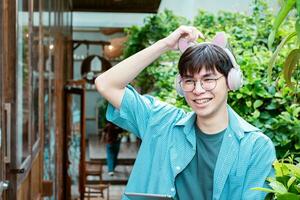 retrato de asiático joven adulto en azul camisa, vistiendo los anteojos, participación auriculares y en pie en frente de el puerta de café comercio, concepto para estilo de vida de adolescentes alrededor el mundo. foto