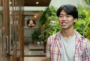 retrato de asiático joven adulto empresario en pie en frente de su propio café tienda cuales situado en rural área, suave y selectivo enfocar. foto