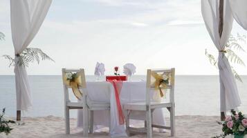 Romantic dinner table set and chair preparation with white theme decoration, white sand, clear cloudy sky, and tranquil, peaceful, beautiful beach view at the seashore. Good place for wedding proposal photo