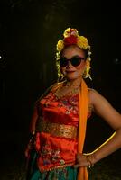 a Sundanese dancer poses in front of a shining light while wearing an orange costume with sunglasses and flowers on her head photo