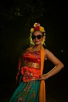 a Sundanese dancer poses in front of a shining light while wearing an orange costume with sunglasses and flowers on her head photo