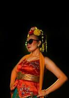 a Sundanese dancer poses in front of a shining light while wearing an orange costume with sunglasses and flowers on her head photo