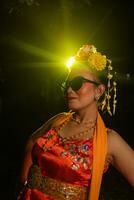 a Sundanese dancer poses in front of a shining light while wearing an orange costume with sunglasses and flowers on her head photo