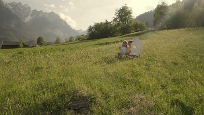 a girl and a boy holding a house cutout sitting in the grass on a