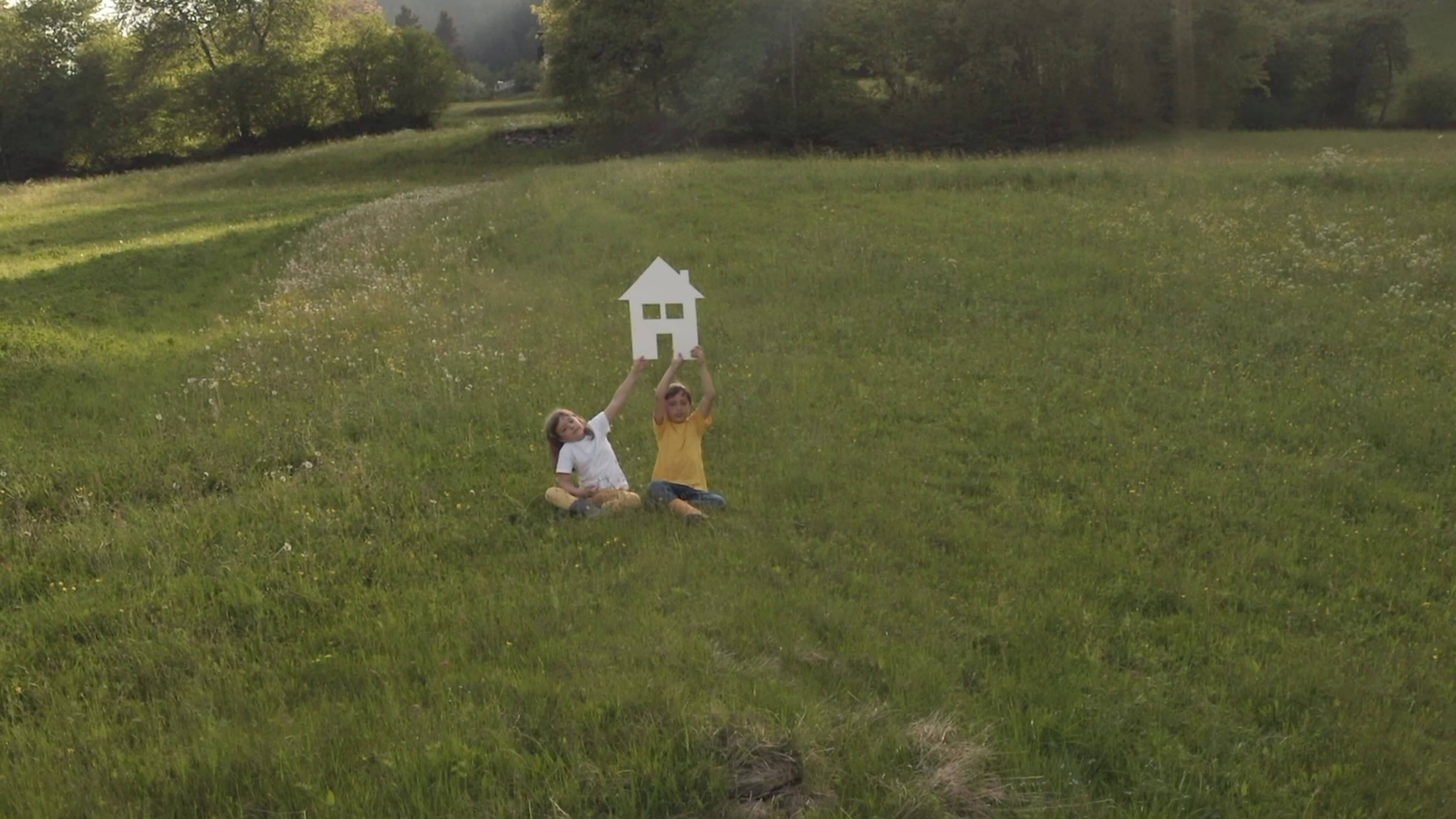 a girl and a boy holding a house cutout sitting in the grass on a