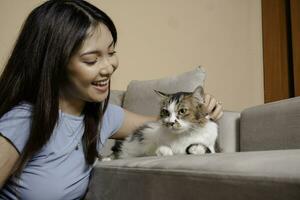 bonito asiático mujer abrazo un gato y sentar en sofá con contento emoción. adorable Doméstico mascota concepto. foto