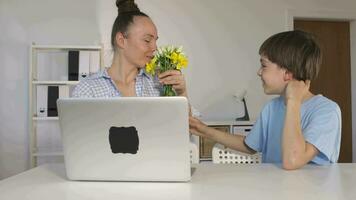 a boy giving a flower to the working mother sitting at a desk with a laptop video