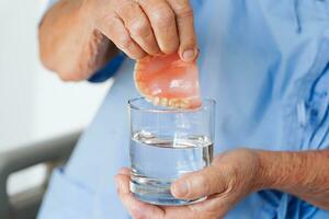 Asian senior woman patient clean teeth denture in a glass with solution for chew food. photo