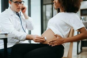 Pregnant african woman has appointment with doctor at clinic. Male gynaecologist OB GYN medic specialist with stethoscope listens to baby's heartbeat in mother's belly. Pregnancy, health care photo