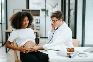 Pregnant african woman has appointment with doctor at clinic. Male gynaecologist OB GYN medic specialist with stethoscope listens to baby's heartbeat in mother's belly. Pregnancy, health care photo