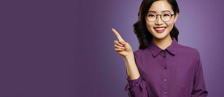 An Asian girl with glasses stands against a purple background smiling and pointing with open palms to advertise while looking joyful photo