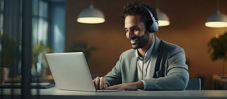 Smiling Middle Eastern male manager in headset working on laptop in modern office consulting clients online using PC photo