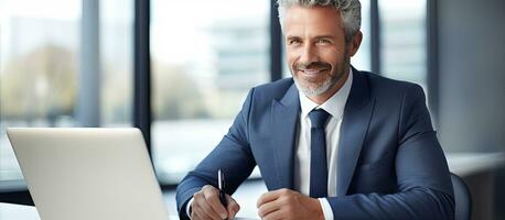 Middle aged professional sitting at desk in office working on laptop and taking notes photo