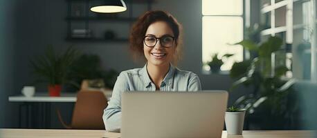 Female entrepreneur working at desk in office smiling at camera with empty laptop screen space for copy photo