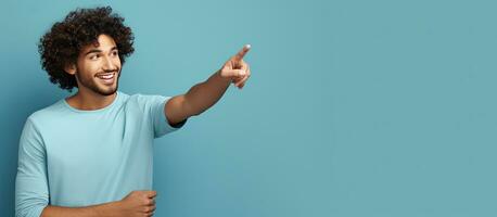 A young Hindu man with curly hair is pointing at a white blank advertising board against a blue background photo