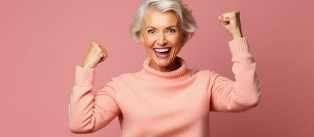 Elderly woman in yellow sweater clenching fists exuding joy isolated on pink background photo