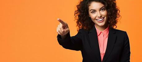 A confident young woman in a black suit and blue blouse is happily pointing at a blank space against a yellow orange background in a wide horizontal im photo