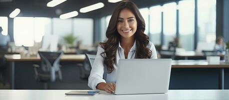 Female entrepreneur working at desk in office smiling at camera with empty laptop screen space for copy photo