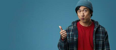 A young Asian man in a beanie and red flannel shirt stands against a blue background pointing up with a puzzled expression photo