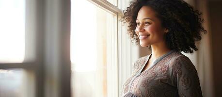 Young pregnant black woman smiling and looking at empty space enjoying her pregnancy near a window at home photo