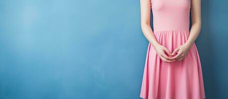 A young woman in a pink dress poses against a blue backdrop displaying a hand with space for text and placing the other hand on her waist photo