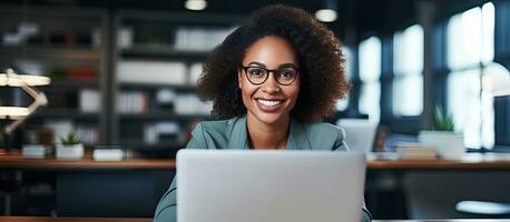 Happy black female executive working on laptop in contemporary office and smiling at camera photo