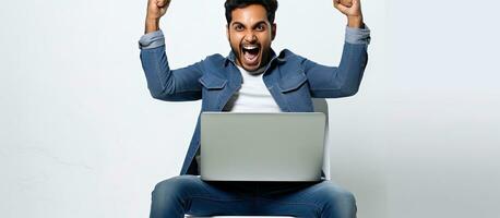 Enthusiastic Indian man celebrating success on laptop sitting on chair against white wall background looking at computer screen and shaking fists happily photo
