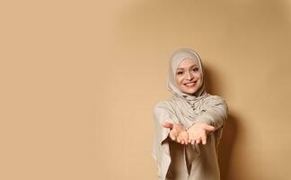 contento musulmán mujer en hijab y estricto atuendo sonrisas lindo y estiramientos fuera su manos a el cámara mientras en pie en un beige antecedentes foto