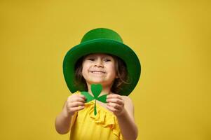 Pretty little girl holds a clover leaf in hands and shows it to camera. Saint Patrick's Day concept on yellow background. photo