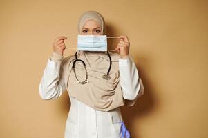 Muslim doctor with covered head in hijab holding and wearing medical mask standing on beige background eith copy space photo