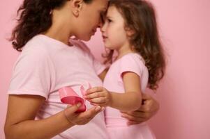 Focus on pink satin ribbons, symbol of International Breast Cancer Awareness Day, on hands of blurred loving mother and lovely daughter background . Female healthcare and medical education concept photo