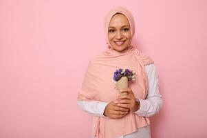 Isolated portrait on colored background of attractive Muslim Arab woman in pink hijab, holding a bouquet of wildflowers in purple shades, wrapped in craft paper, smiles at camera with a toothy smile photo