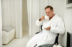 Handsome mature 50 years old man, successful businessman in white bathrobe drinking tea from a cup while relaxing in privacy room in luxury wellness spa center. Male beauty revitalizing treatment photo