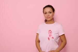 confidente retrato de un sereno joven mezclado carrera africano mujer vistiendo un rosado camiseta con rosado satín cinta en símbolo de internacional día de cáncer conciencia. oncológico enfermedades concepto. Copiar espacio foto