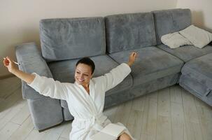 Smiling woman sits on the floor in the hallway of her apartment and stretches her arms to the sides looking towards the light from the window photo