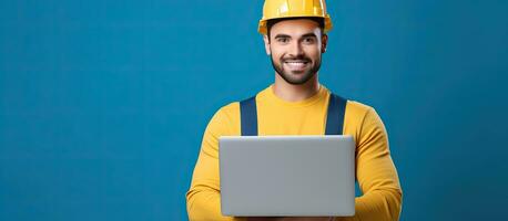 Young man in protective headgear and attire holds laptop smiles at camera Isolated on yellow background Copy space photo