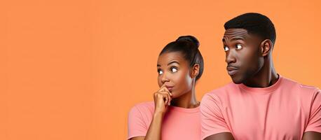 Thoughtful African couple in orange shirts directing attention to blank area isolated on pink backdrop photo