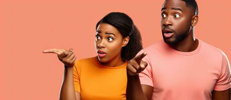 Thoughtful African couple in orange shirts directing attention to blank area isolated on pink backdrop photo