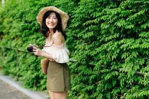 Portrait of asian young woman traveler with weaving hat and basket and a camera on green public park nature background. Journey trip lifestyle, world travel explorer or Asia summer tourism concept. photo