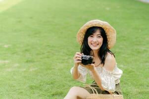 retrato de asiático joven mujer viajero con Costura sombrero y cesta y un cámara en verde público parque naturaleza antecedentes. viaje viaje estilo de vida, mundo viaje explorador o Asia verano turismo concepto. foto