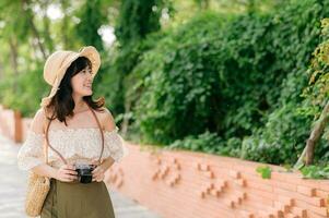 Portrait of asian young woman traveler with weaving hat and basket and a camera on green public park nature background. Journey trip lifestyle, world travel explorer or Asia summer tourism concept. photo