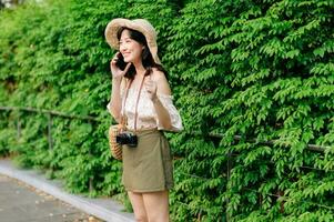 Portrait of asian young woman traveler with weaving hat, basket, mobile phone and camera on green public park background. Journey trip lifestyle, world travel explorer or Asia summer tourism concept. photo